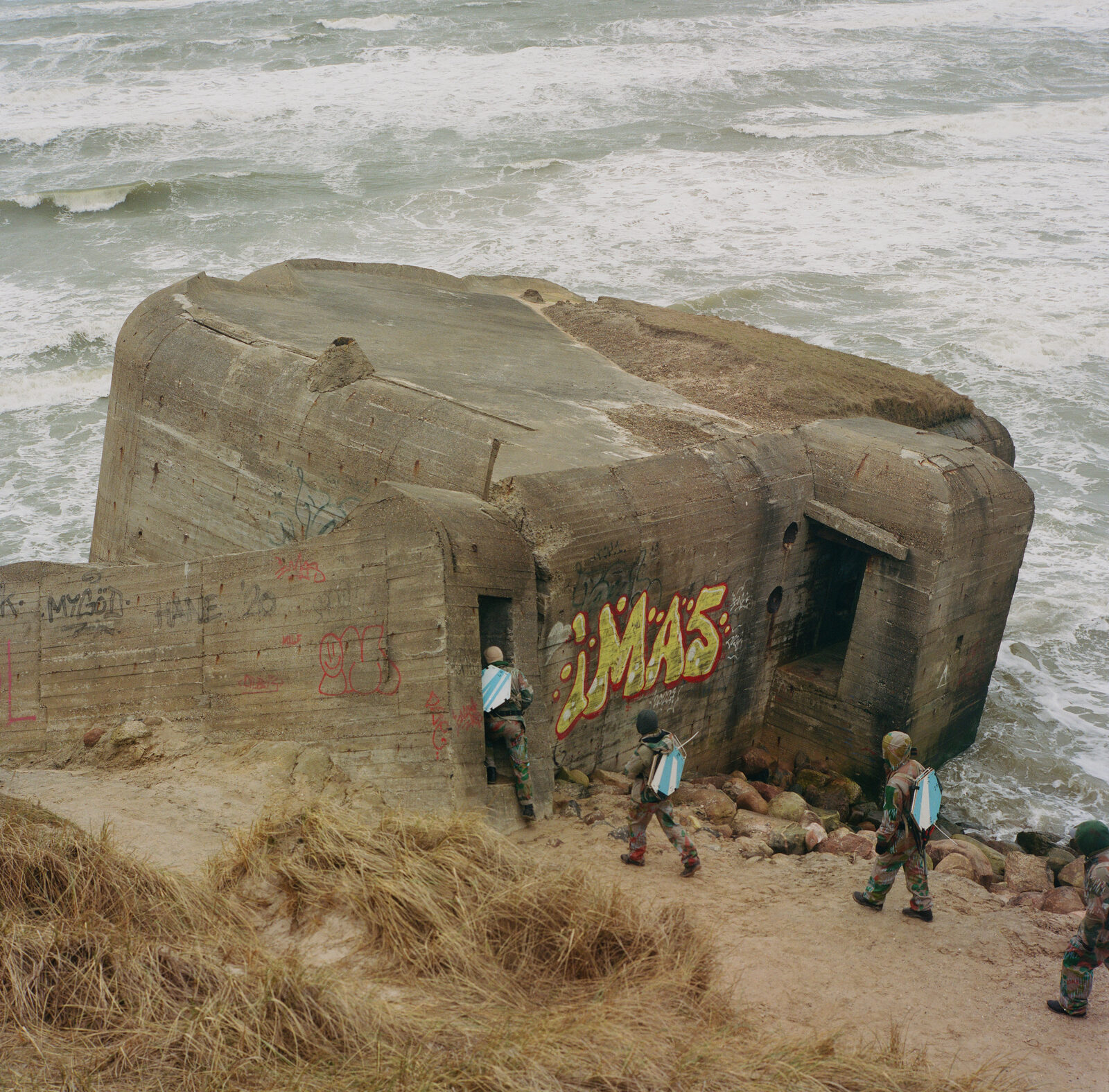 Figures in printed uniform entering a WWII bunker on a stormy coastline