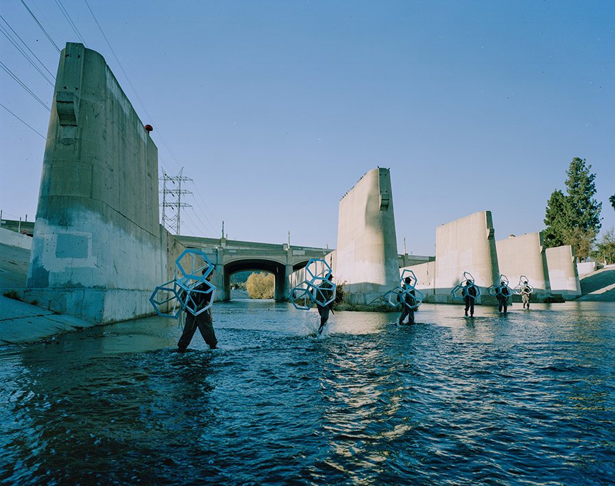 Figures carrying wireframe hexagonal forms crossing the Los Angeles River at Glendale